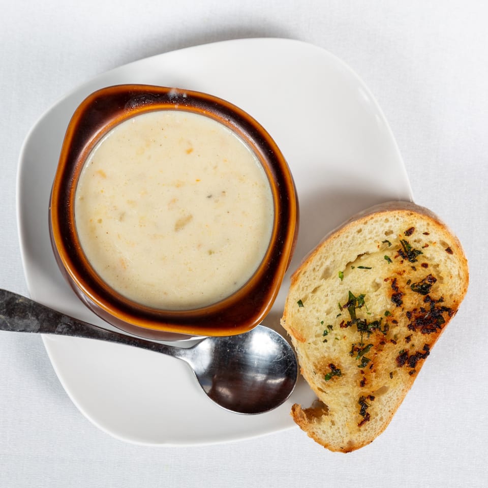 Creamy soup in a brown bowl with a spoon and a slice of toasted garlic bread on a white plate.