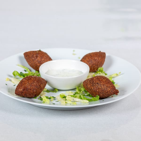 Four kibbeh, football-shaped fried croquettes, with a white dipping sauce and lettuce on a white plate.