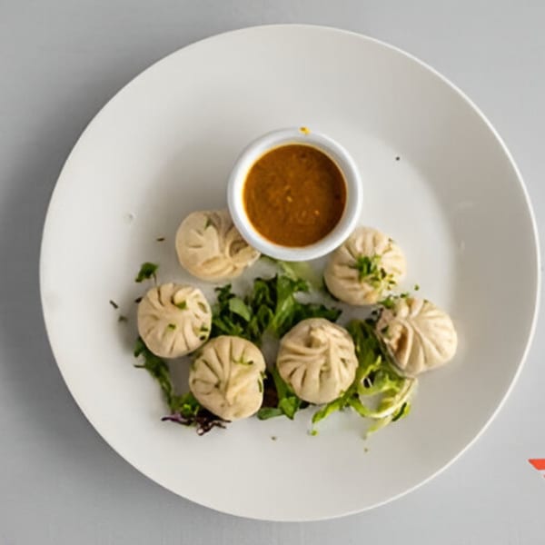 Six steamed dumplings with green garnish and a side of dipping sauce on a white plate.