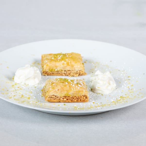 Two pieces of baklava with whipped cream, powdered sugar, and pistachio dust on a white plate.