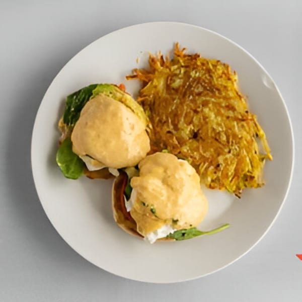Two eggs benedict with hollandaise sauce and spinach, served with a side of hash browns.