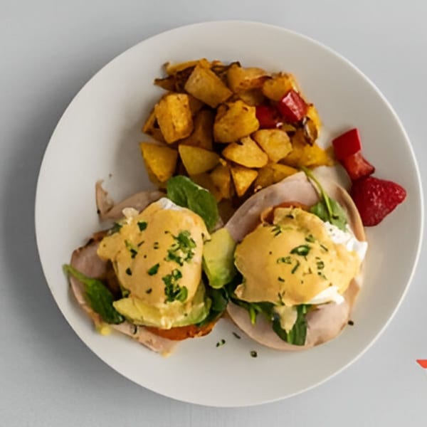 Two eggs benedict with avocado, spinach, and turkey, served with roasted potatoes and red peppers.