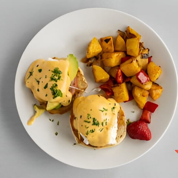 Eggs Benedict with avocado and bacon, served with roasted potatoes and strawberries on a white plate.