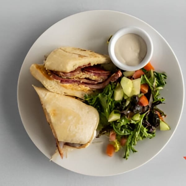 A Lighthouse Bistro sandwich with meat and cheese, served with a side salad and dipping sauce.