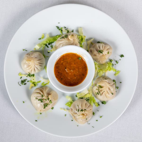 Six steamed dumplings with a side of spicy sauce and lettuce on a white plate.