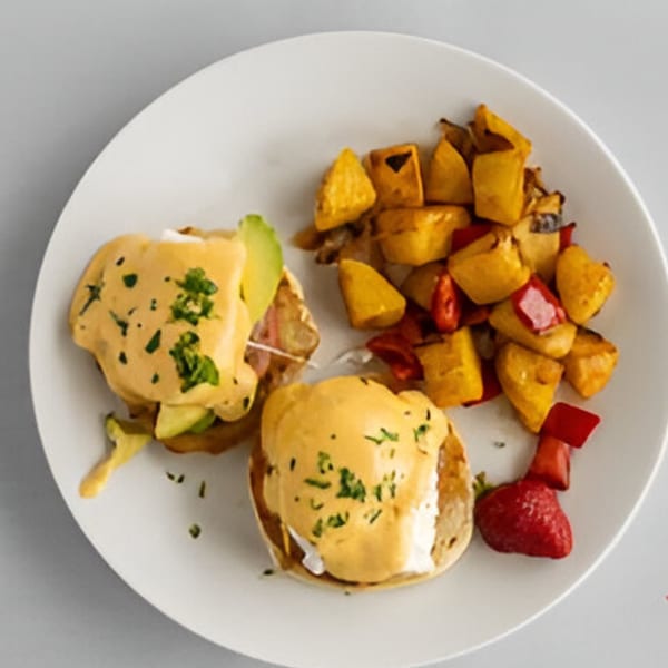 Two eggs benedict with avocado, roasted potatoes, and red peppers on a white plate.